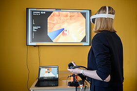 Annika Köhler ist von hinten zu sehen, sie trägt eine VR-Brille und hält das Endoskop in der Hand, an der Wand ist die virtuelle Welt einer endoskopischen Behandlung projiziert. 