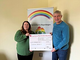 Auf dem Foto sieht man zwei Personen, die symbolisch einen großen Scheck von 2.000 Euro in der Hand halten. Im Hintergrund steht ein Roll-up von der Elterninitiative Regenbogen.