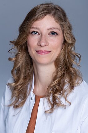 Porträtbild von Carolin Julia Curtaz im Hochformat - die Ärztin in weißem Kittel hat dunkelblonde, lange, gelockte Haare und blaue Augen