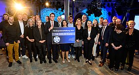 Gruppenbild mit mehreren Personen. Prof. von Oertzen und Frau Nelkenstock halten einen Spendenscheck auf dem Förderpreisverleihung 2025 und 16.500 Euro steht. Auf dem Scheck ist links das Logo Forschung hilft zu sehen. Der Hintergrund leuchtet blau und an der Decke hängen viele Scheinwerfer. Links leuchtet einer der Scheinwerfer direkt in die Kamera.