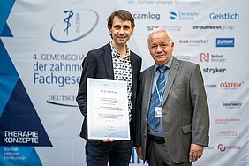 Ralf Krug und Edgar Schäfer stehen vor einer Logo-Wand - Ralf Krug hält die Urkunden in der rechten Hand. 