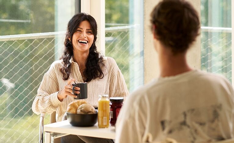 Person sitzt an einem Frühstückstisch, lächelt in die Kamera und hält einen Kaffee in der Hand