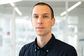 Porträt von Jonas Czwikla im Foyer des Deutschen Zentrums für Herzinsuffizienz - Czwikla trägt sehr kurze, dunkle Haare und ein dunkles Hemd 