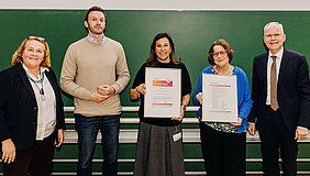 Auf dem Bild stehen drei Damen und zwei Herrn bei der Urkundenübergabe vor einer grünen Tafel. Zwei der Damen halten eine Urkunde in der Hand.