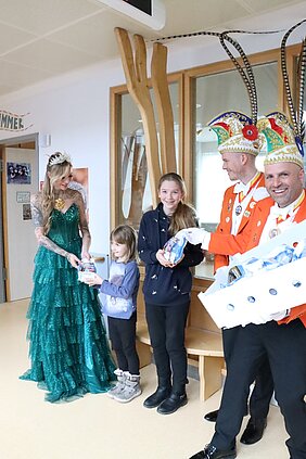 Prinzessin Katharina im langen grünen Kleid überreicht Krapfentüten an zwei Kinder, rechts im Bild 2 Männer vom Elferrat in Faschingskluft mit orangefarbenen Jackets und langen Federn am Hut