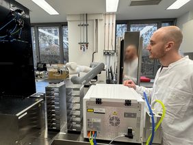 Frederik Erkens steht im weißen Kittel neben der Anlage und schaut auf den Roboterarm, sein Profil spiegelt sich im schmalen Kühschrank. 