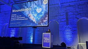 Vanessa Borst steht auf der Bühne hinter dem Rednerpult - im Hintergrund ist der Monitor mit ihrer Präsentation Wound Ambit - die Bühne  erscheint in blauem Licht 