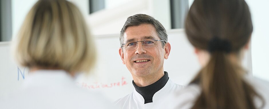 Prof. Dr. Stefan Störk Stefan Störk im weißen Kittel steht vor HI-Nurses, ebenfalls in weißer Arbeitskluft, im Hintergrund ein Flipchart