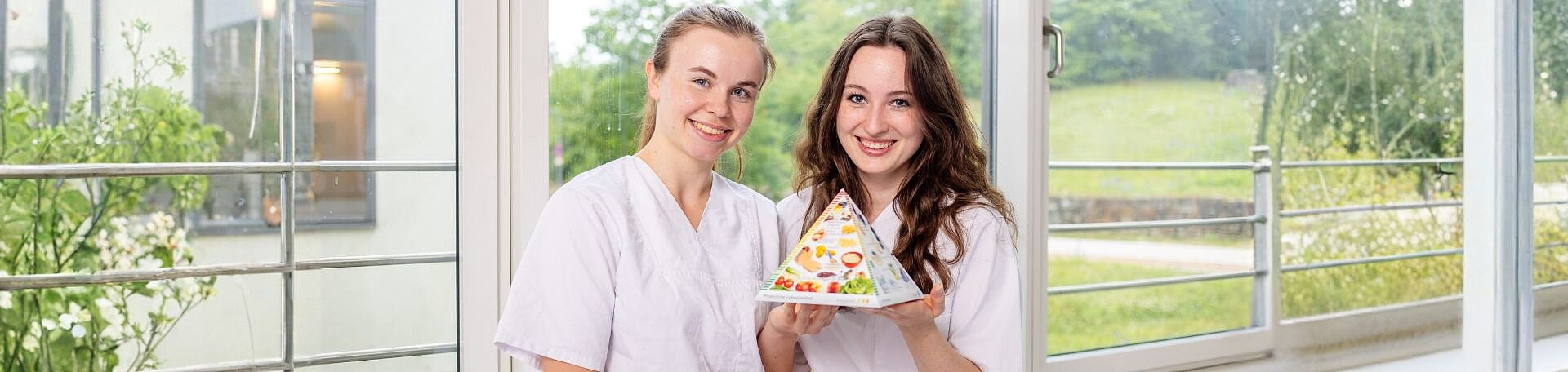 Welche Rolle spielt die Ernährung bei der Behandlung von Erkrankungen? Das wissen Diätassistentinnen und Diätassistenten genau. Zwei Ausbildende der Diätassistenz halten das Modell einer Ernährungspyramide hoch.