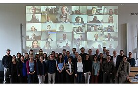 Gruppenfoto nach Verabschiedung der Satzung. Im Hintergrund sind an einer Wand die virtuellen Teilnehmer zu sehen. 
