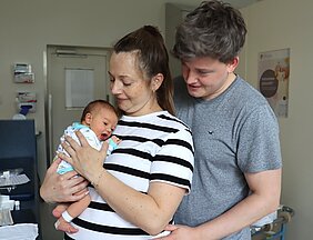 Die Mutter hält den Säugling auf dem Arm, der Vater steht daneben. 