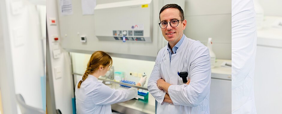 Charis Kalogirou mit weißem Kittel im Labor - im Hintergrund arbeitet eine Mitarbeiterin an der Laborbank. 