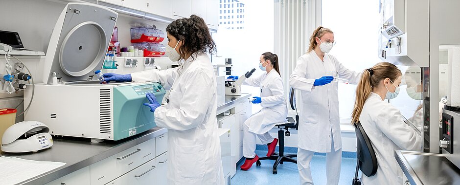 4 Mitarbeiterinnen der Nuklearmedizin am DZHI im Labor an verschiedenen Geräten, alle mit Mundschutz und weißen Kitteln. 