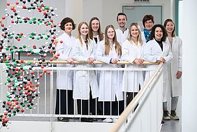 Die neun Mitarbeitende des Referenzdiagnostik-Labors stehen in weißen Kitteln im Foyer des Biozentrums, daneben hängt ein DNA-Mobile. 