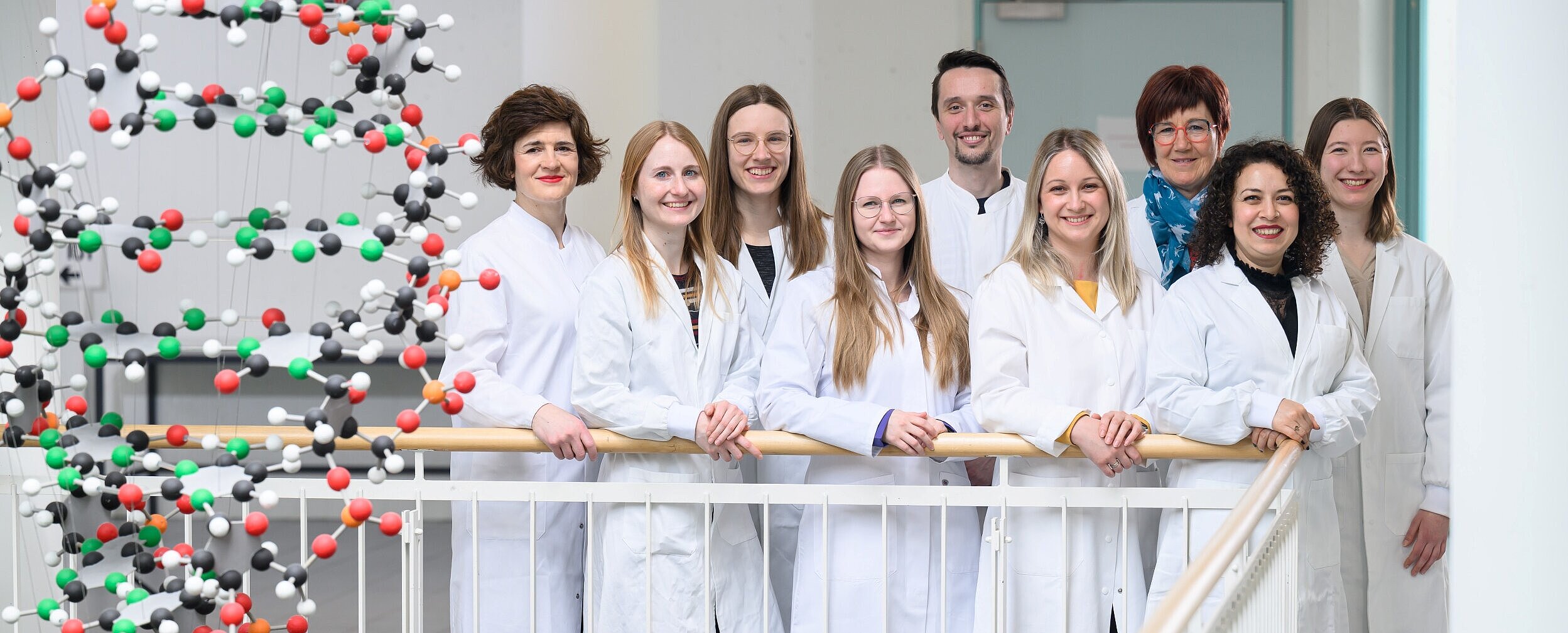 Die neun Mitarbeitende des Referenzdiagnostik-Labors stehen in weißen Kitteln im Foyer des Biozentrums, daneben hängt ein DNA-Mobile. 