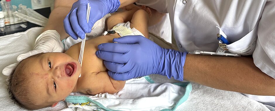 Säugling liegt auf Wickelkommode, Studienärztin nimmt mit Wattepad einen Abstrich von der Haut, im Hintergrund steht die Studynurse mit Mundschutz