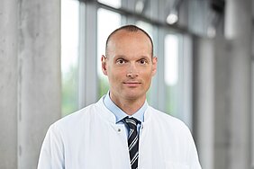 Prof. Dr. Rafael Jakubietz steht in der Magistrale des ZIM/ZOM. Er trägt eine schwarz/weiße Krawatte und ein blaues Hemd welches aber durch den weißen Arztkittel größtenteils verdeckt wird. Im Hintergrund erkennt man verschwommen zwei graue Betonpfosten und dazwischen eine helle Fensterfront. 