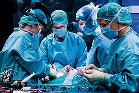 4 Fachkräfte in OP-Kluft mit Kittel, Haube und Mundschutz stehen am Operationstisch und behandeln einen Patienten