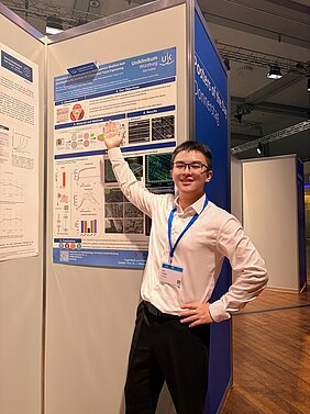 Zhi Liang präsentiert sein Wissenschaftsposter auf dem DOG-Kongress - er steht vor dem Ausdruck an der Posterwand und hält Arm in die Höhe 