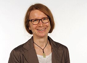 Porträt von Barbara Namer, die einen braunen Blazer trägt, eine Lederkette mit Anhänger, die braunen Haare locker zusammengebunden. 