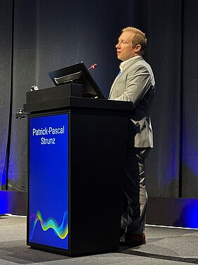 Patrick-Pascal Strunz steht am Podium auf der Bühne vom EULAR-Kongress in Barcelona.