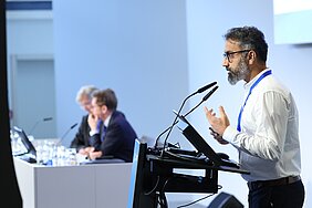 Malik Salman Haider steht am Rednerpult und spricht ins Mikro, das Bild ist von der Seite aufgenommen worden. 