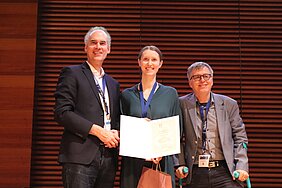 Preisträgerin mit Urkunde auf der Bühne, links steht Martin Fassnacht, rechts von ihr Jan P. Tuckermann mit Krücken. 