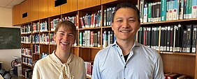 Lisa Harder-Rauschenberger und Chi Wang Ip stehen in der Bibliothek der Neurologie vor einem Bücherregal und schauen in die Kamera. 