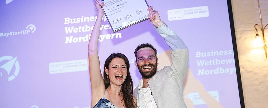 Anna Fleischer im blau-weißen Sommerkleid und Johannes Braig im beigefarbenen Jacket halten auf der Bühne die  eingerahmte Urkunde hoch und strahlen in die Kamera. 