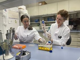 Die beiden Wissenschaftlerinnen sitzen mit weißen Kitteln im Labor und pipettieren.