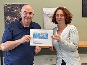 Auf dem Bild sind zwei Personen zu sehen, die in der Hand eine Karte halten auf denen verschiedene rote Punkte zu sehen sind. Über der Karte steht Bavaria-Queensland Research Alliance. Im Hintergrunde hängen zwei Bilder an der Wand.