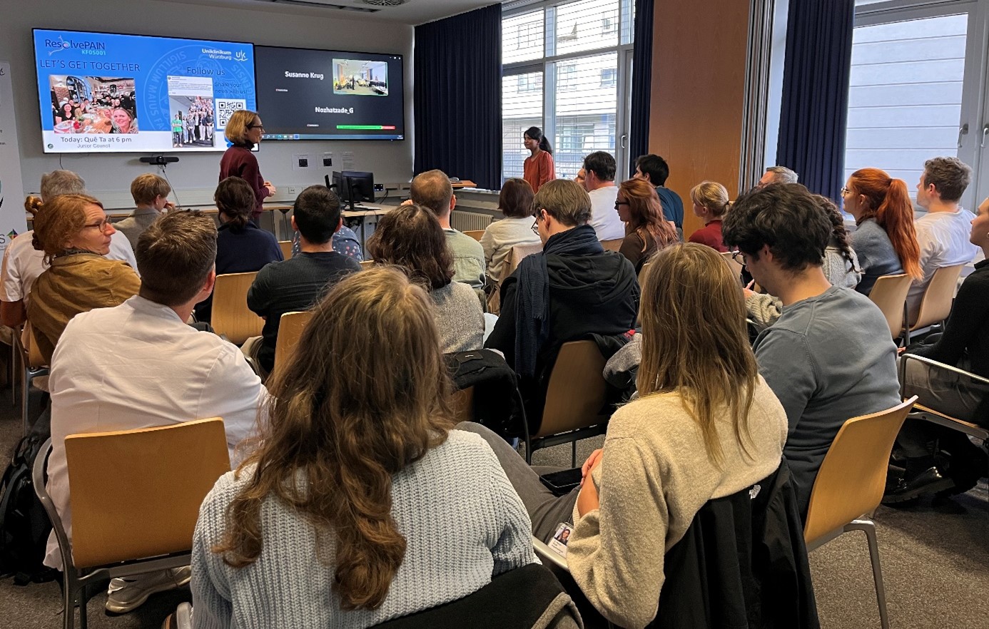 Prof. Dr. med. Heike Rittner und Dr. Ankita Rawat präsentieren ihre Forschungsergebnisse. Auf einem großen Bildschirm an der Wand läuft eine Powerpoint-Präsentation. Etwa 25 Zuhörerinnen und Zuhörer folgen dem Vortrag.