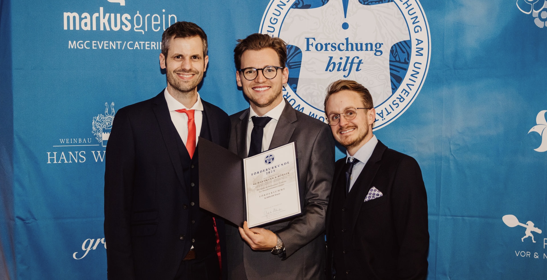 Die Preisträger PD Dr. med. Dr. med. dent. Stefan Hartmann und Dr. med. dent. Marius Hörner vor dem Logo der Stiftung "Forschung hilft". Marius Hörner hält die Förderurkunde in Händen.