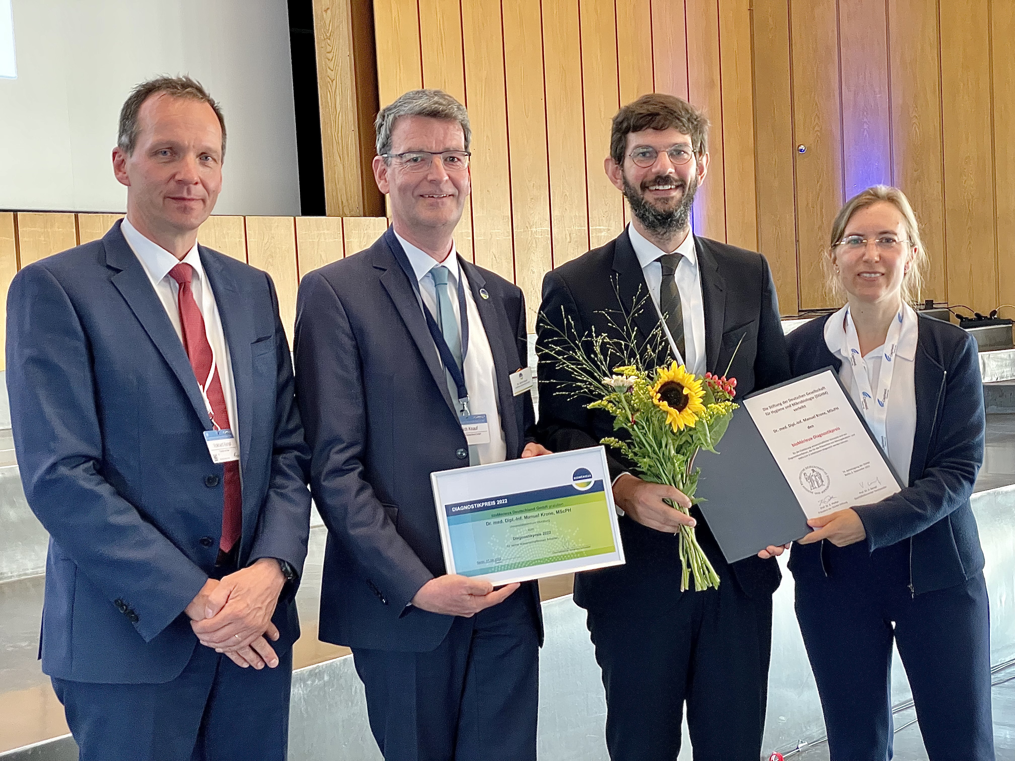 Das Bild zeigt Manuel Krone bei der Preisverleihung mit Blumen, Scheck und Urkunde.