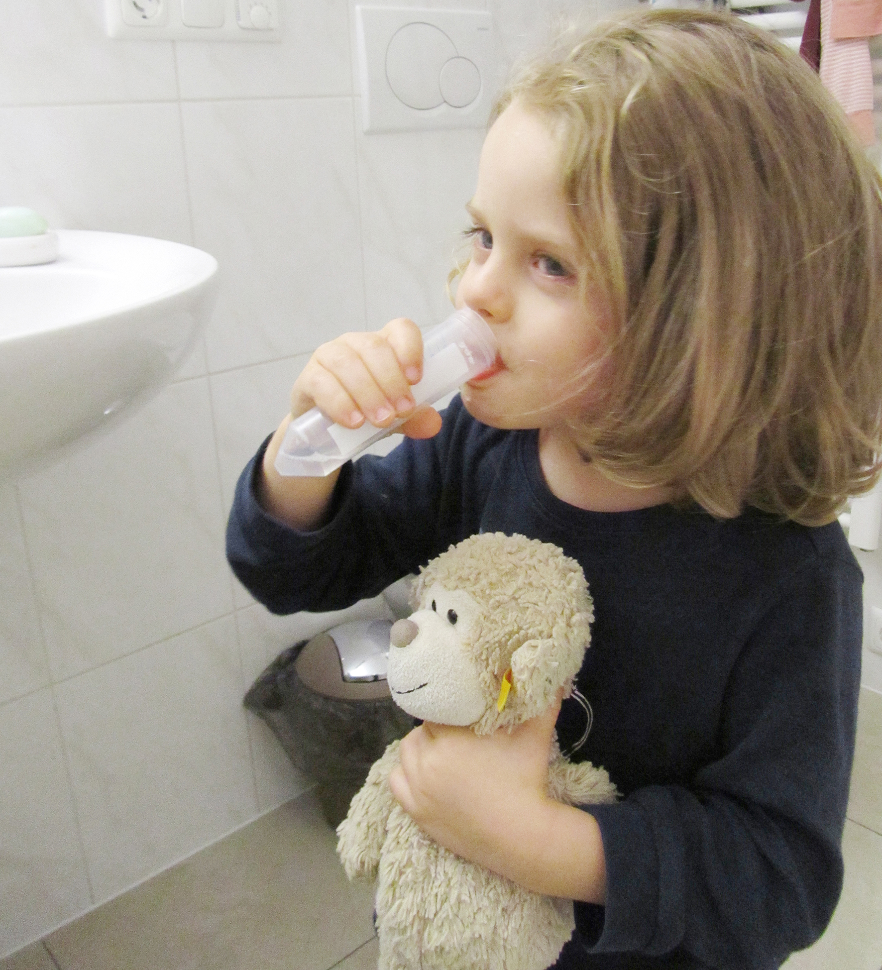 Das Bild zeigt ein Mädchen, das in einer Hand ihren Teddy hält und in der anderen einen Probebehälter aus dem es gerade trinkt. 