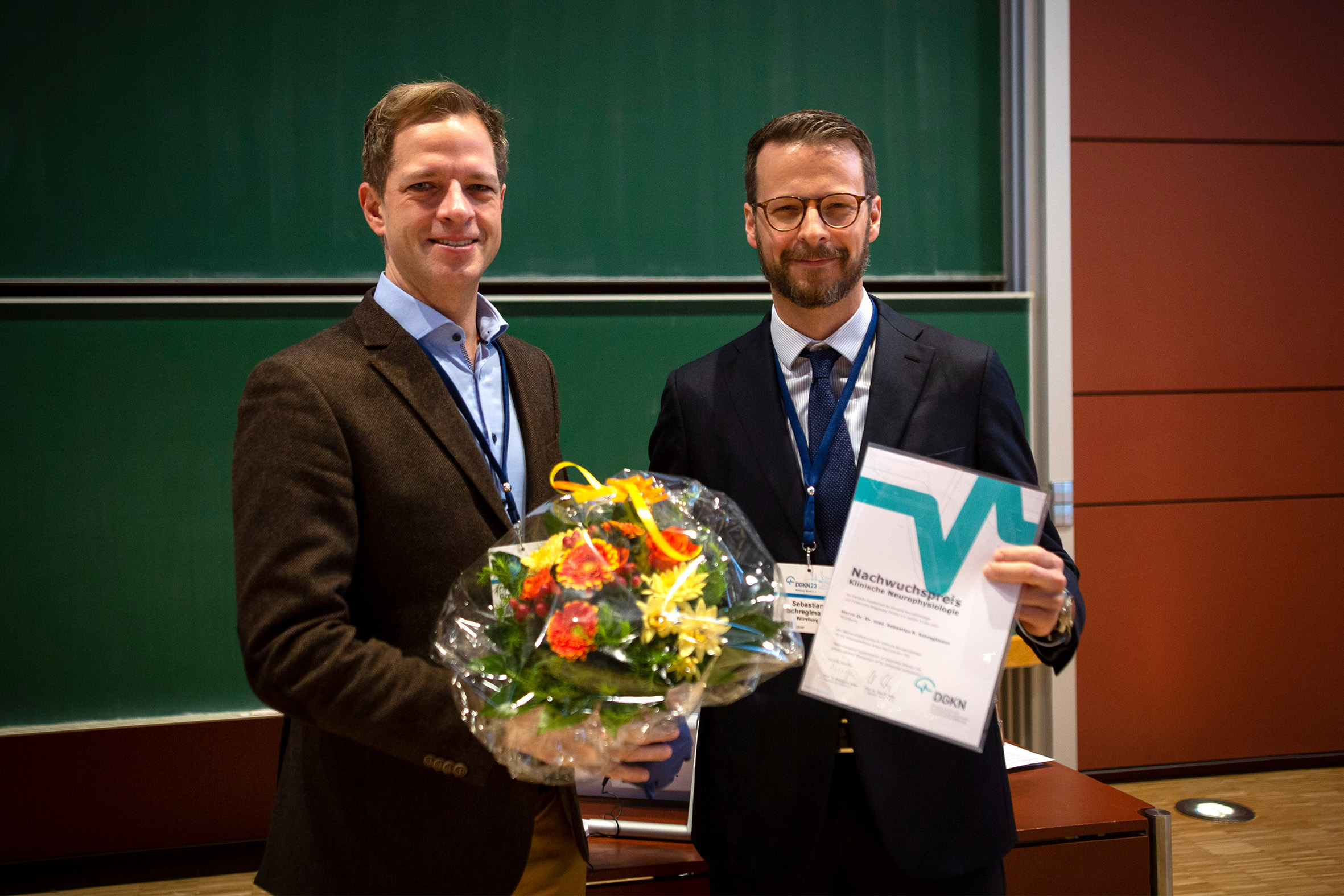 Links im Bild ist Prof. Christian Grefkes-Hermann mit Blumenstrauß, rechts Dr. Sebastian Schreglmann, der die Urkunde in den Händen hält. 