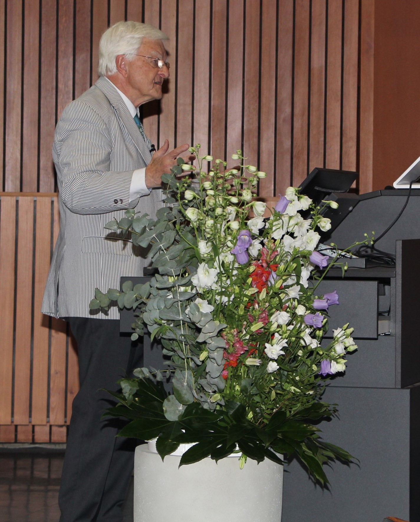 Klaus Viktor Toyka steht am Rednerpult im Hörsaal, vor ihm ein großer bunter Blumenstrauße in einer Bodenvase. 