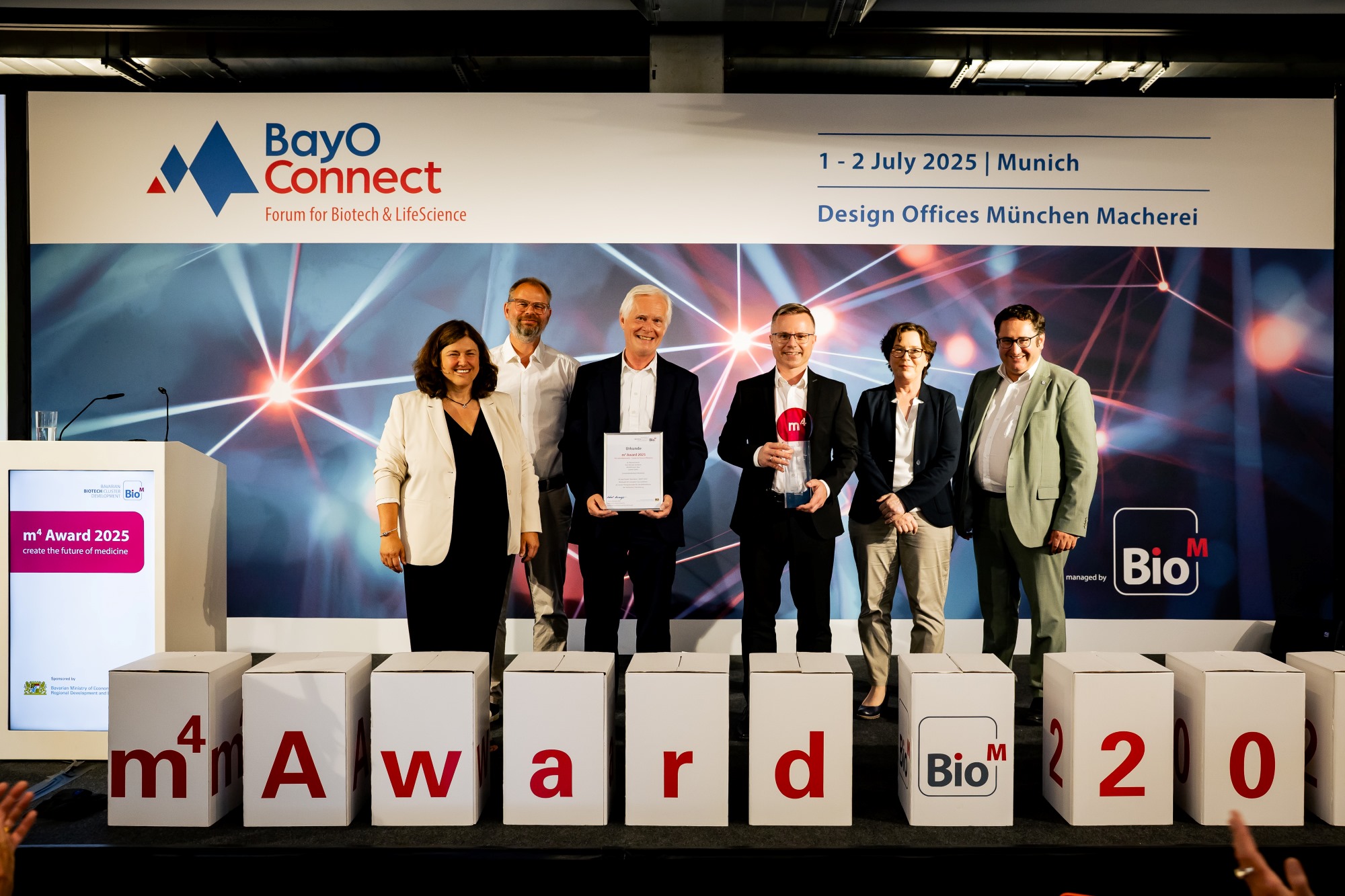 Die Preisträger mit Organisatorinnen und Staatssekretär auf der Bühne - davor sind Würfel mit Buchstaben m4 award