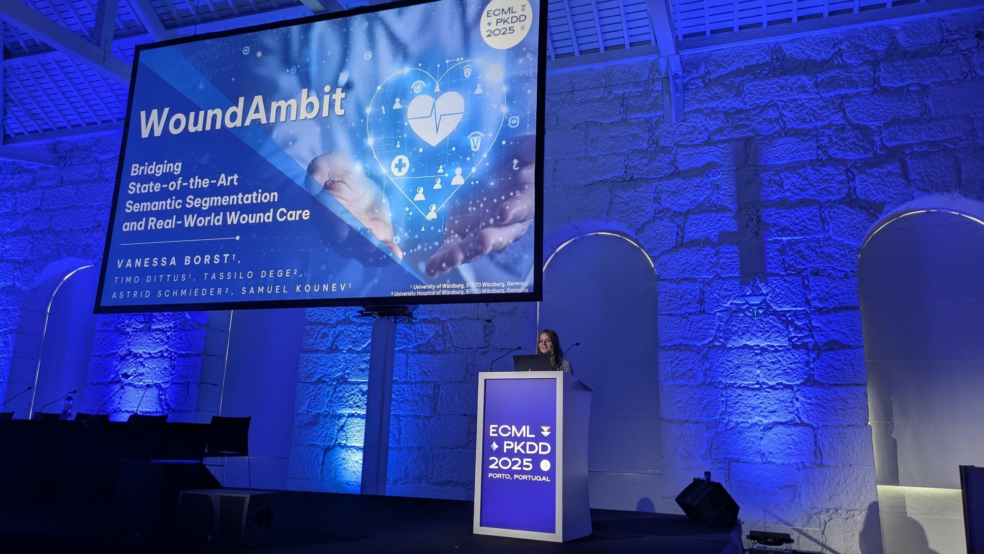 Vanessa Borst steht auf der Bühne hinter dem Rednerpult - im Hintergrund ist der Monitor mit ihrer Präsentation Wound Ambit - die Bühne  erscheint in blauem Licht 