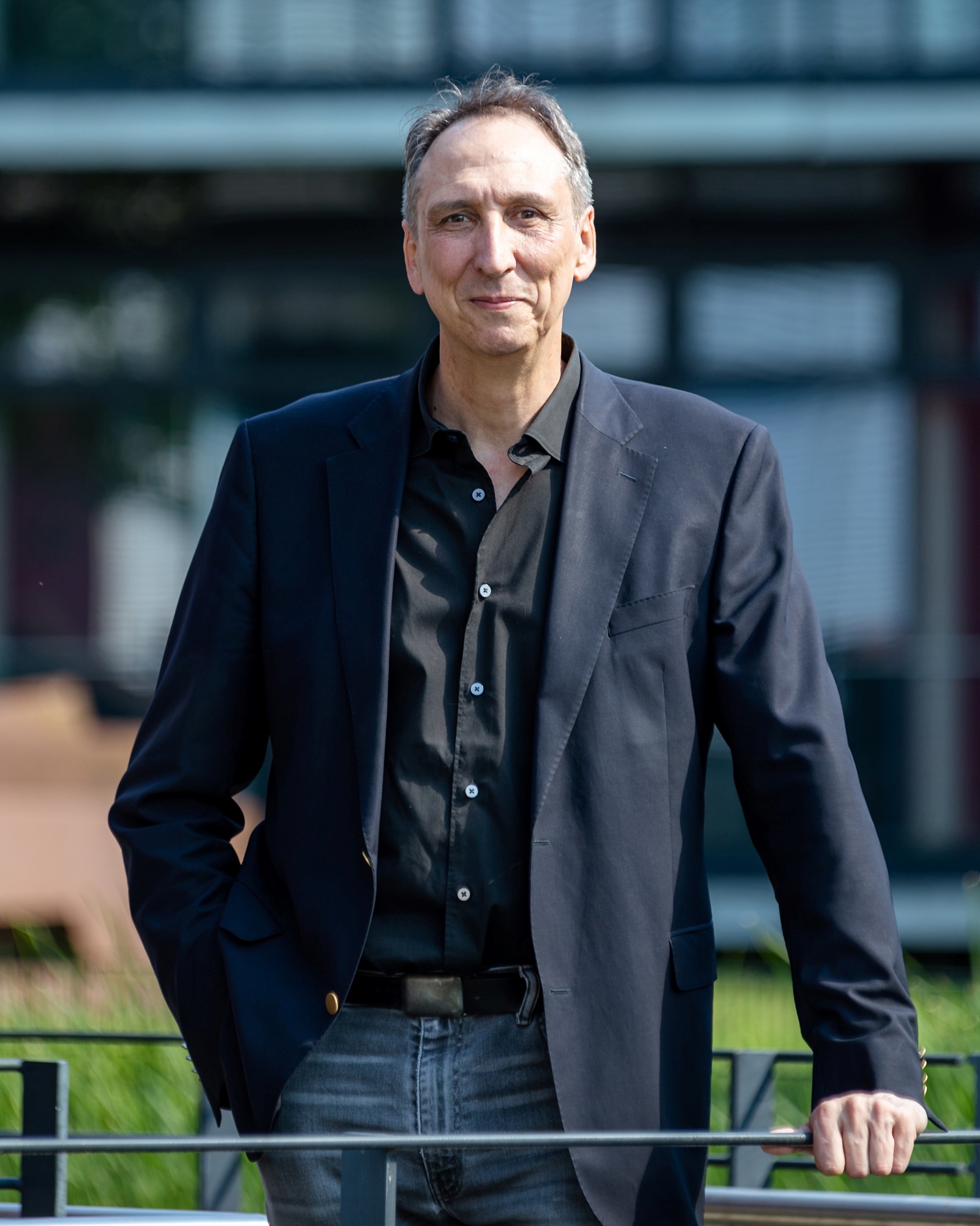 Prof. Dr. Meining steht schmunzelnd hinter einem Geländer an welchem er sich mit seiner linken Hand festhält. Im Hintergrund kann man eine Wiese und ein verglastes Gebäude, welches aber verschwommen dargestellt wird, erkennen.