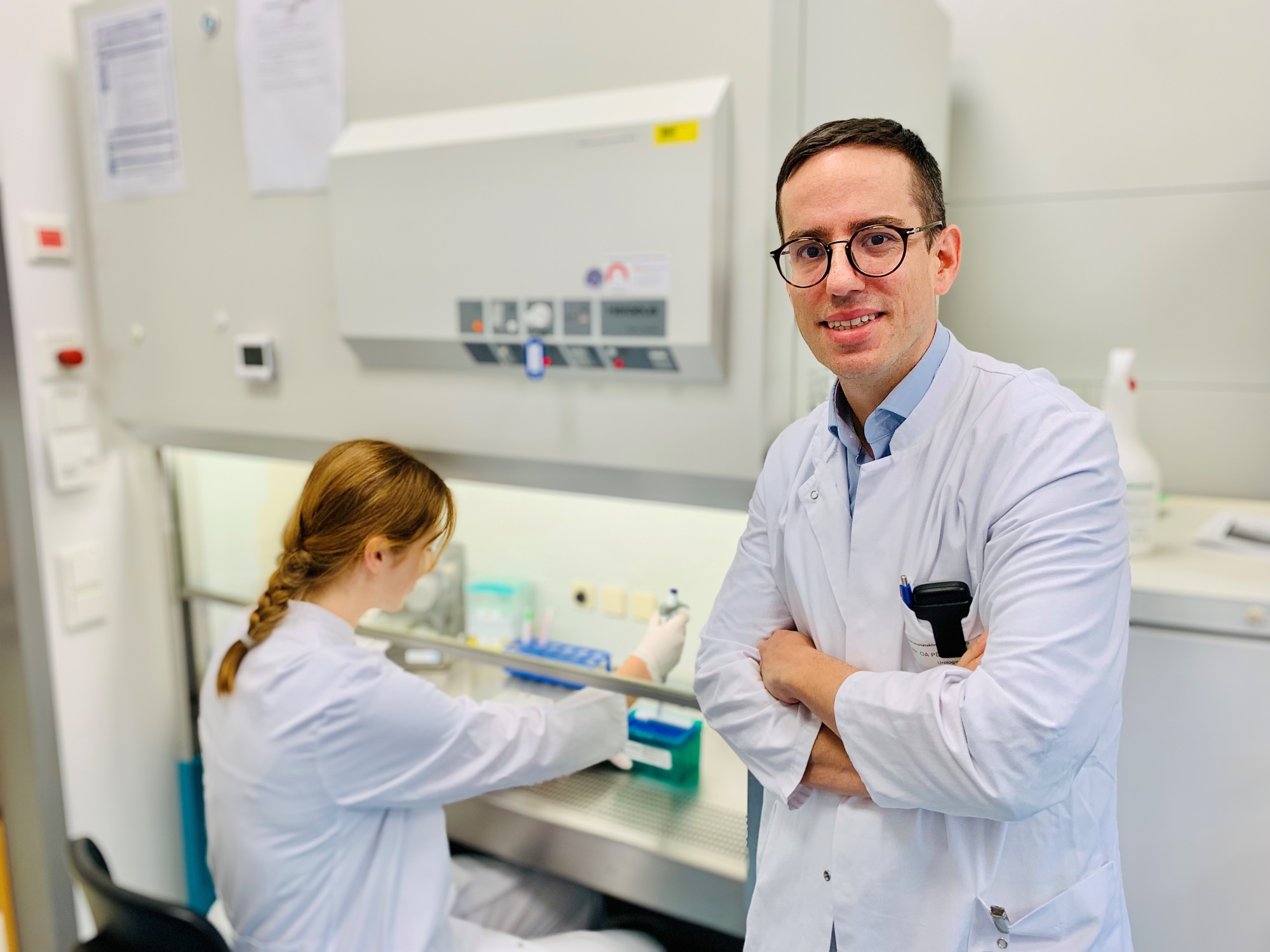 Charis Kalogirou - Urologie Der Oberarzt steht im Labor, er trägt einen weißen Kittel, seine Arme sind verschränkt, im Hintergrund ist eine Mitarbeiterin zu sehen, die an einer Laborbank arbeitet.