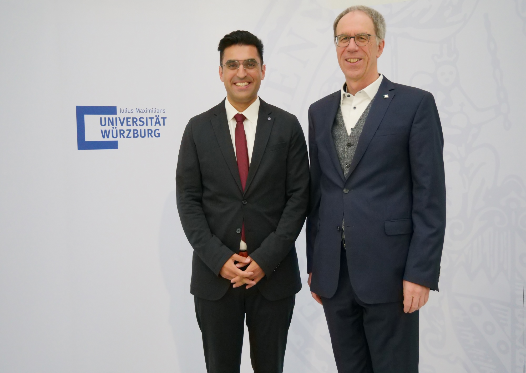Ugur Uslu und Paul Pauli posieren in Anzügen nebeneinander vor einer Wand mit Logo der Universität Würzburg 