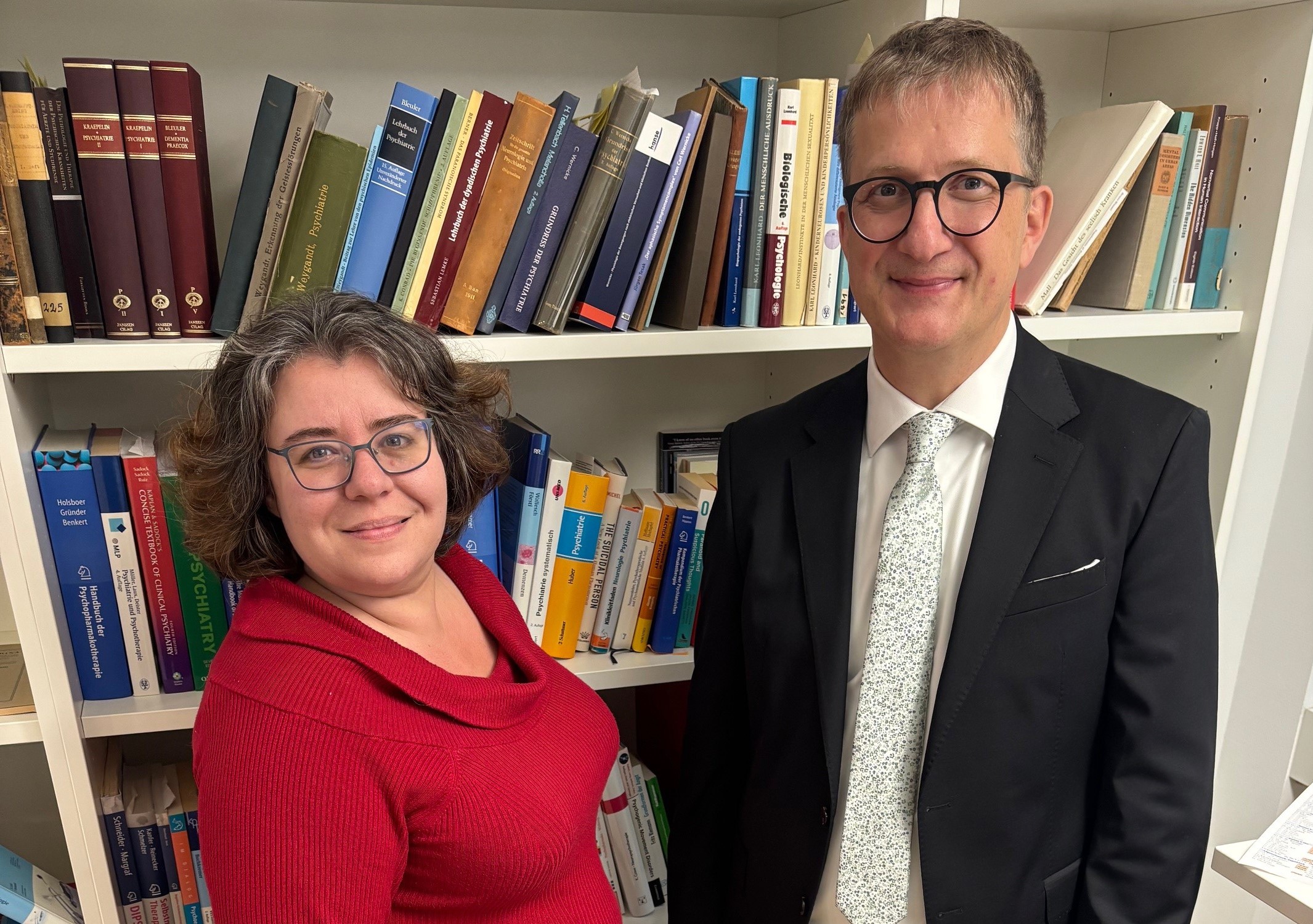 Die Wissenschaftler Stephanie Lefebvre und Sebastian Walther stehen vor einem Bücherregal, Lefebvre hat braune gewellte Haare und einen roten Rollkragenpullover, Sebastian Walther trägt Anzug und helle Krawatte.