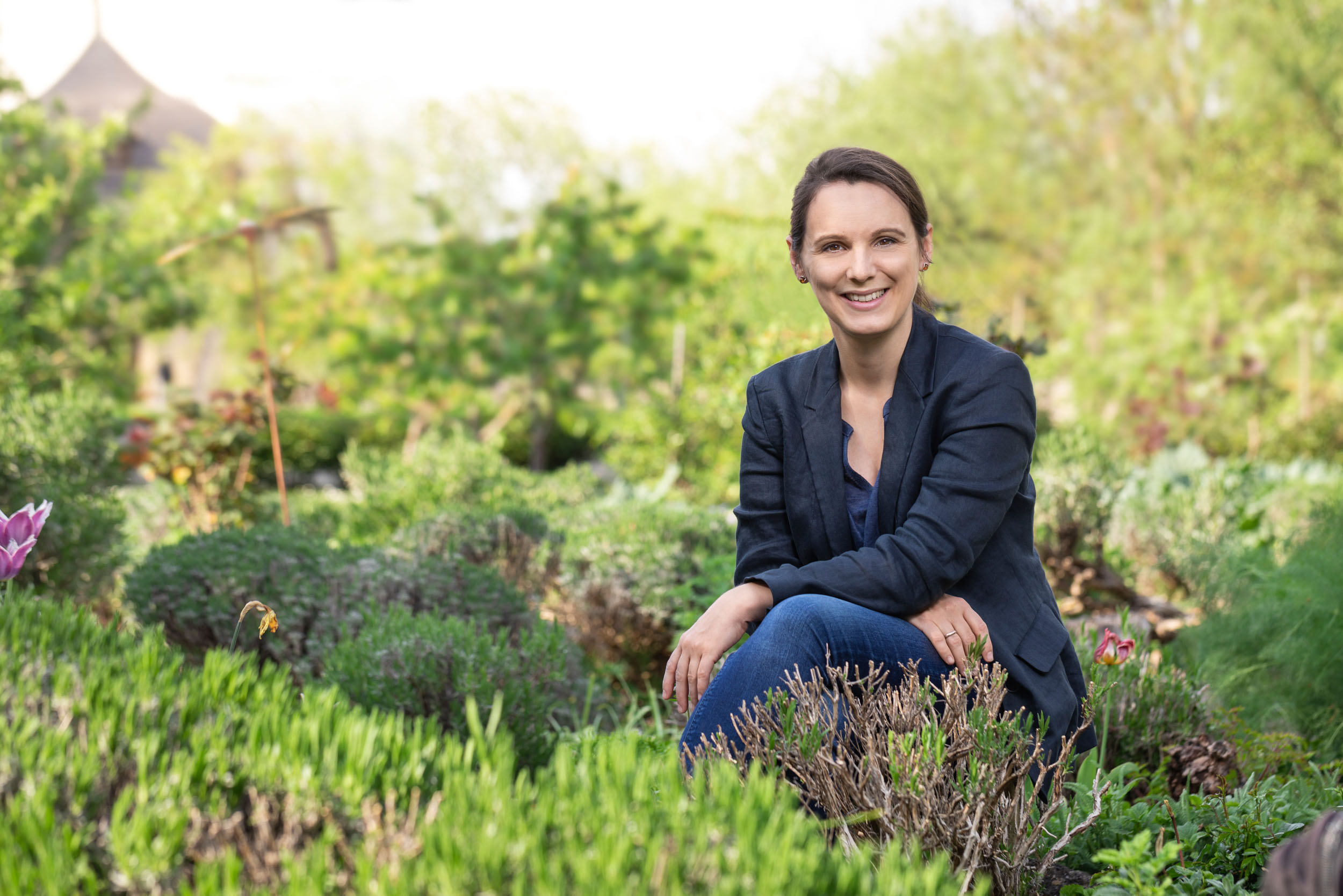 Claudia Löffler mit Jeans und dunklem Blazer kniet in einem liebevoll angelegten Garten 