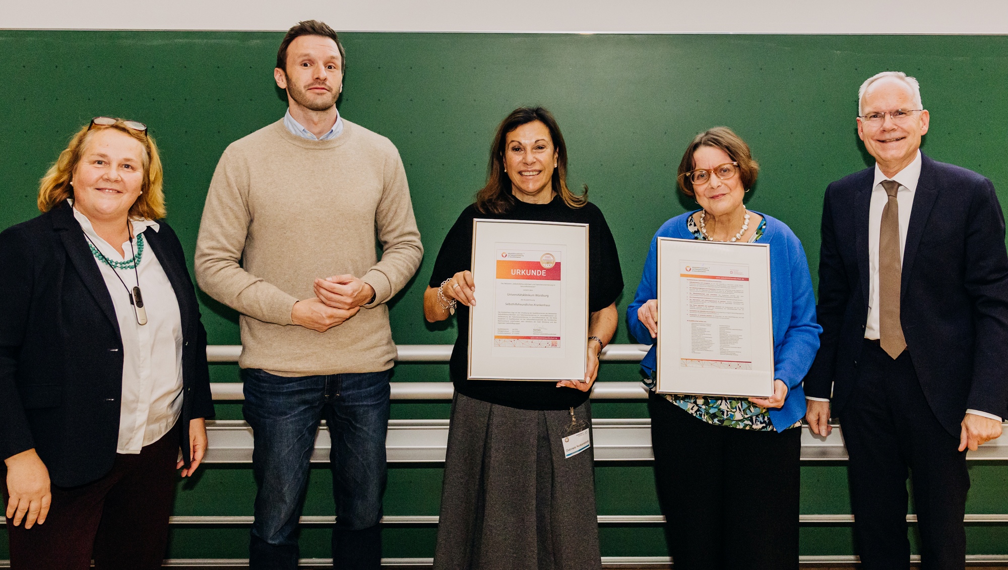 Auf dem Bild stehen drei Damen und zwei Herrn bei der Urkundenübergabe vor einer grünen Tafel. Zwei der Damen halten eine Urkunde in der Hand.