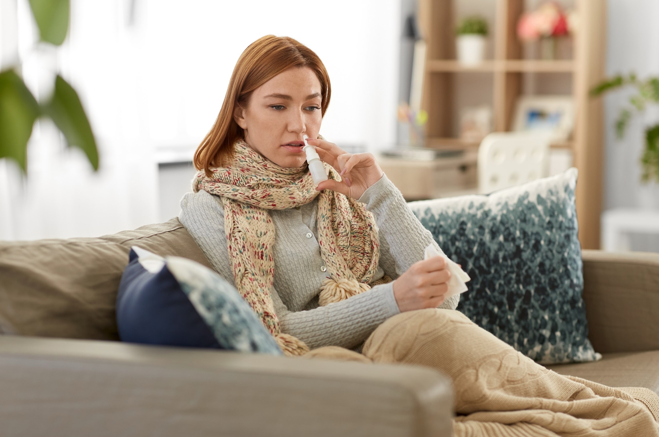 Frau mit Schal und Decke sitzt auf einem Sofa - mit einem Taschentuch in der einen Hand und einem Nasenspray in der anderen Hand. Das Nasenspray führt sie gerade zur Nase. 