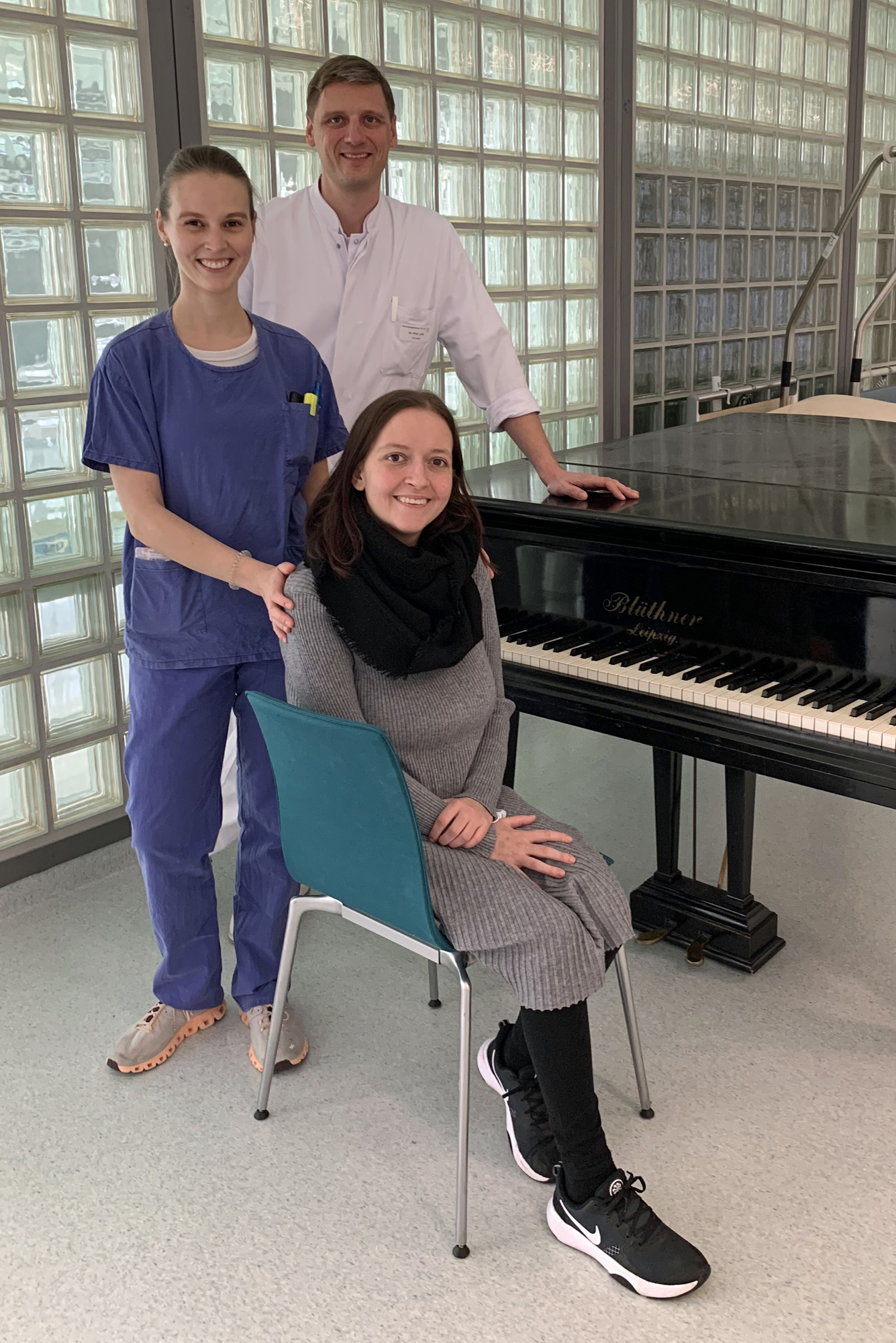 Auf dem Bild sind drei Personen zu sehen. Im Hintergrund ist eine Glassteinwand zu sehen. Die Ärztin hat blaue OP-Kleidung an und hat ihre Hände auf den Schultern der Patientin. Der Prof. hat einen weißen Kittel an und stützt sich mit einer Hand auf einem Klavier ab. Die Patienten sitzt auf einem Stuhl vor dem Klavier.