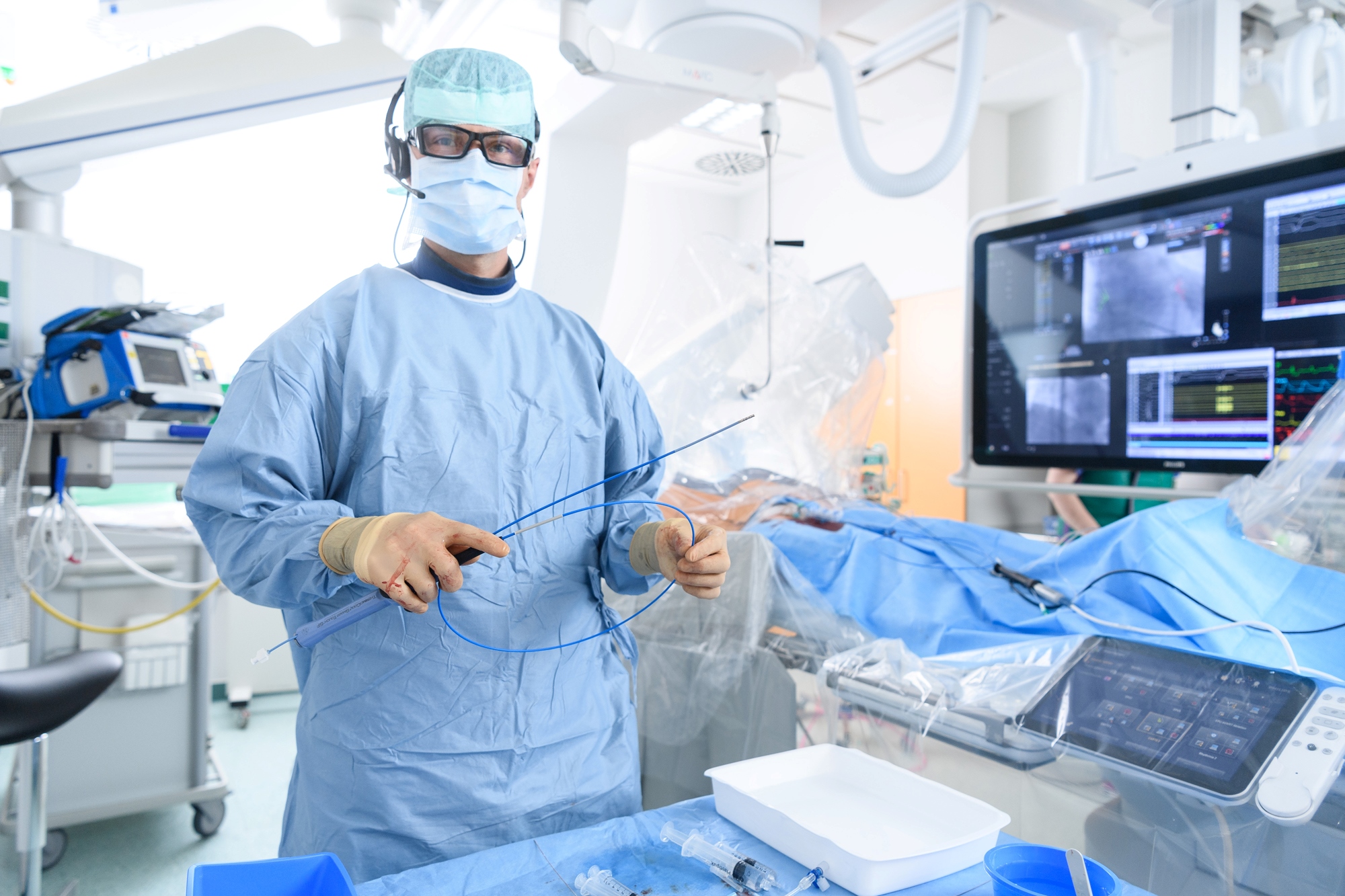 Das Bild zeigt einen OP-Saal. Im Hintergrund sieht man einen Monitor und mehrere OP-Geräte. Vor ihm steht ein Tisch mit Spritzen und seitlich eine OP-Liege. Ein Arzt hält in der Hand einen Herzkatheter. Er trägt eine Brille und einen Mundschutz, Handschuhe, eine Kopfbedeckung sowie einen blauen OP-Kittel. Zusätzlich trägt er auch noch ein Headset. 