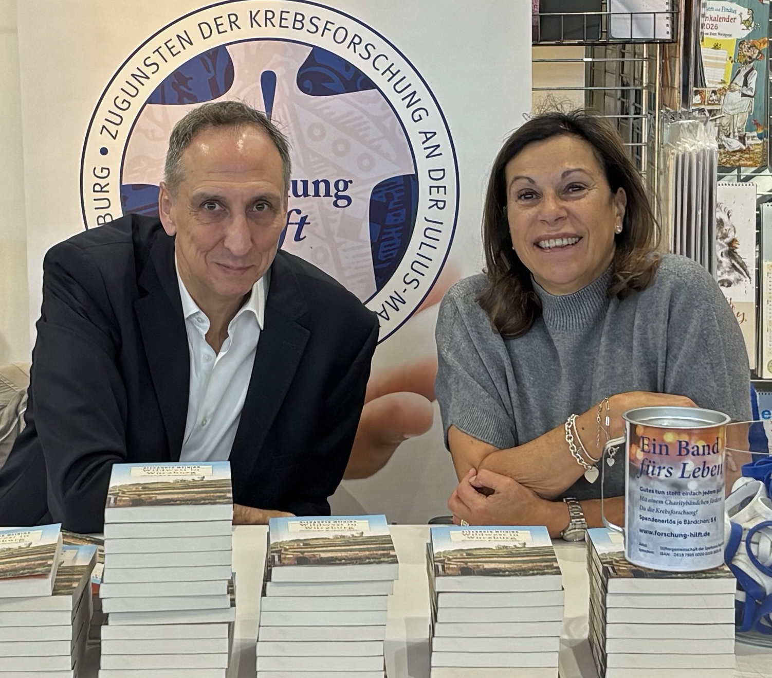 Prof. Meining und Frau Nelkenstock sitzen an einem Tisch. Davor liegen mehrere Stapel Bücher. Auf einem der Stapel steht eine Spendenbox. Im Hintergrund steht ein Rollup. Auf dem ein Kreis mit dem Text Zugunsten der Krebsforschung zu sehen ist. 
