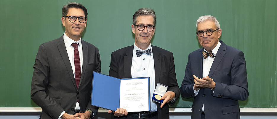 Auf dem Bild sind drei Herren zu sehen, die vor einer grüner Tafel stehen.  Die eine Person in der Mitte hält eine Urkunde und eine Medaille in der Hand. Die rechte Person klatscht in die Hände. 